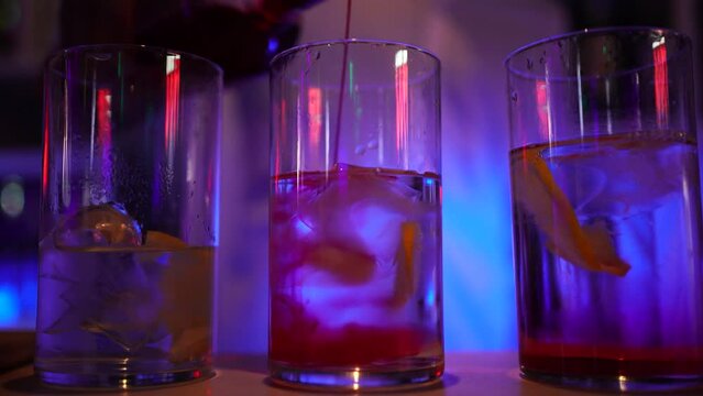 Tres copas de alcohol y refresco con hielos y fruta encima de una barra de discoteca en orden ascendente. La acción se hecha granadina y se ve cómo se difumina en color rojo. 