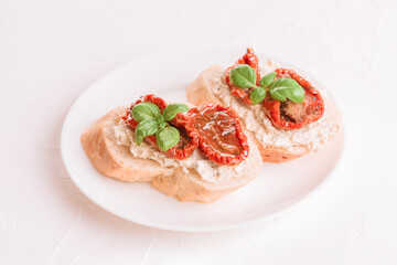 Bruschettas or toasts with ricotta cheese and sliced sun dried tomatoes topped with basil on white plate, white table background. Delicious italian appetizer, antipasti
