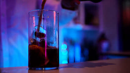 Aparece una copa de alcohol con refresco oscuro, se ve cómo cae el refresco sobre los hielos y la fruta. En el fondo el camarero con las luces de neón de la barra desenfocado.