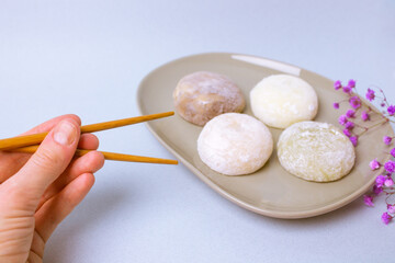 Chopsticks in a hand of a person going to eat a traditional Japanese dessert mochi in rice dough or daifuku. Mochi ice cream balls on gray plate on blue background. Purple flowers decorating a dish.