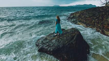 Obraz premium Sexy hot woman meditates, relaxes on a rock reef hill in stormy morning rain cloudy sea. Girl in blue swimsuit, dress tunic. Concept feminine, relax, sexual health. Dark dramatic silhouette view