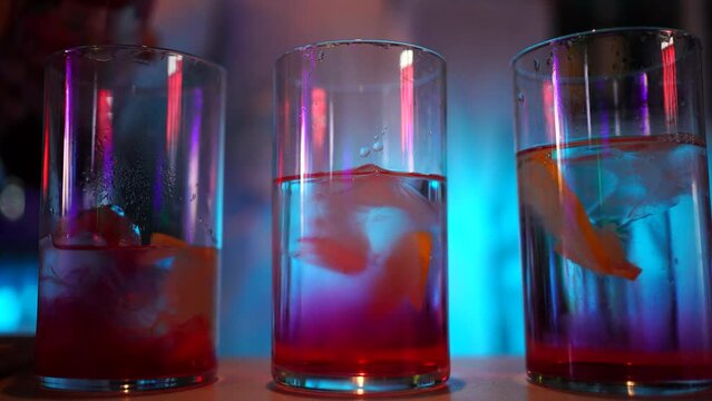 Se sirven 3 copas con hielo y lim&oacute;n en una discoteca encima de una barra con luces de ne&oacute;n. Se aprecia c&oacute;mo cae la granadina en la ginebra provocando un color a rojo. El fondo est&aacute; difuminado