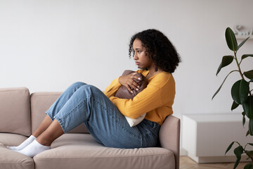 Unhappy young black woman suffering from depression, hugging pillow, sitting on couch at home