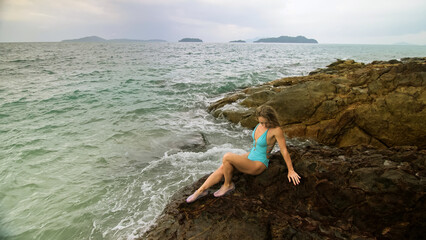 Handsome long-haired woman chill relax resting on rock of sea reef stone, stormy cloudy ocean. Woman in blue swimsuit dress tunic. Concept rest tropical resort coastline traveling outdoor tourism