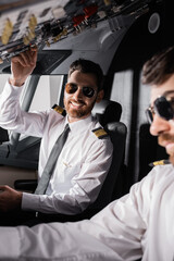 cheerful pilot in sunglasses reaching overhead panel near co-pilot in airplane simulator.