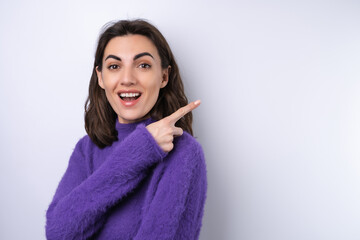 Young woman in a purple soft cozy sweater on a background of cute smiling cheerfully, in high spirits, points a finger to the right to an empty space.