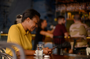African american woman using a smartphone and drinking coffee while sitting in a cafe