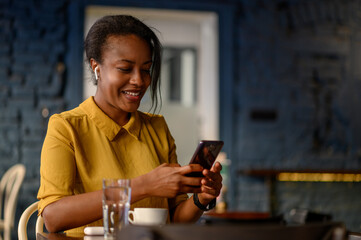 African american woman using airpods and a smartphone while sitting in a cafe