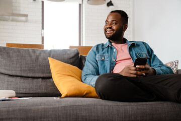 Black bristle man smiling and using cellphone while sitting on sofa