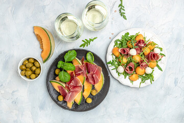 mediterranean salad with prosciutto or jamon, mozzarella and green basil leaves and Cantaloupe melon on white table, traditional Spanish and Italian appetizer served with wholemeal grissini, top view