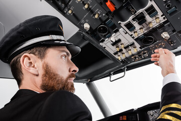bearded pilot in cap reaching overhead panel with set of switches in airplane simulator.