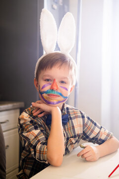 A boy in a white bunny ears headband with a face painted with paint looks at the camera. Fun and Easter