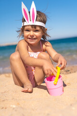 Little girl in swimsuit and bunny hairband sitting on the sandy beach with coloful eggs. Happy...