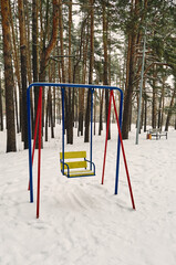 In a public park there are metal swings for entertainment and recreation for children, tall pine trees are all around, a lot of snow has fallen, a winter view