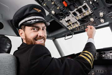 cheerful pilot in cap reaching overhead panel with set of switches in airplane simulator.