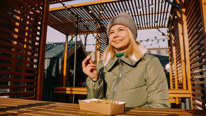 Woman eating tasty french fries in outdoor cafe. Sunny winter day. Woman at an urban outside street food court.