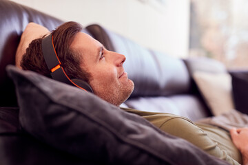 Man Wearing Wireless Headphones Listening To Music Lying On Sofa At Home Wearing Pyjamas