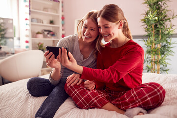 Obraz premium Mother With Teenage Daughter Wearing Pyjamas Sitting On Bed Posing For Selfie On Mobile Phone