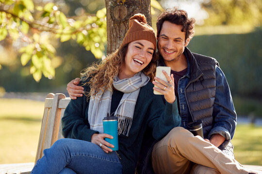 Smiling Couple Sitting On Bench In Autumn Park Using Mobile Phone
