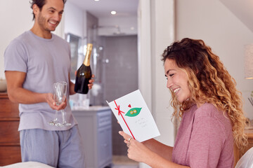 Couple In Bedroom As Man Holds Champagne As Woman Reads Card Celebrating Birthday Or Anniversary