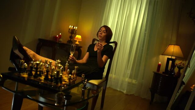 Careless beautiful redhead woman smoking cigar drinking wine sitting with feet on table looking at chess board. Wide shot angle view of relaxed Caucasian chess player indoors