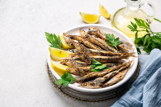 Pan-fried anchovies in the white plate with lemon and parsley. White background, blue runner. Copy space.