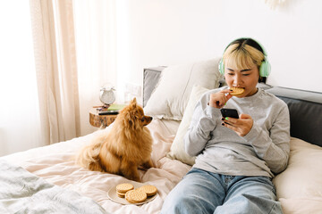 Asian girl eating cookie and using cellphone while resting on bed