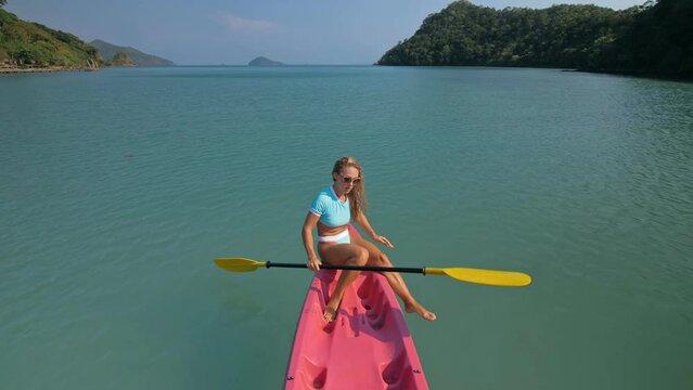 Pretty sportive blonde lady in stylish swimsuit sails on pink plastic canoe along azure ocean bay at exotic resort. Traveling to tropical countries. View from above.