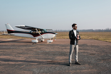 full length of stylish pilot in leather jacket and sunglasses standing near helicopter.