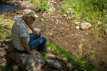 Old man with hat sitting meditates in front of a stream.