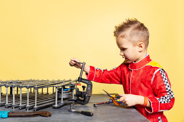 Cute little boy, child in image of auto mechanic or fitter in dungarees with work tools repairing auto motor isolated on yellow background.