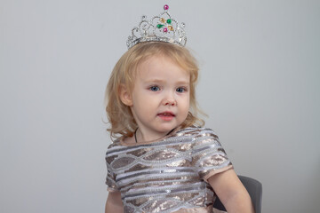 Fototapeta na wymiar A beautiful baby girl 2 years old in a silver crown and a shiny dress is sitting on a chair on a gray background