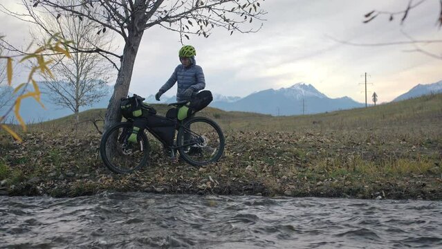 The woman travel on mixed terrain cycle touring with bikepacking. The traveler journey with bicycle bags. Sportswear in green black colors. The trip in magical autumn forest, river stream.