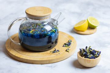 A glass teapot made of organic blue anchana on a wooden stand and a cup of dry, blue pea tea flowers with lemon slices on a light table. Herbal tea. Selective focus.