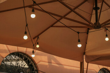 Switched on bulb lights decor hanging across the cafe umbrella of the restaurant terrace. Interior design. Light design
