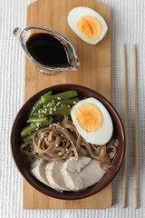 Asian food ramen noodles with meat, vegetables and egg in a brown bowl on a light background, soy sauce in a gravy boat, chopsticks