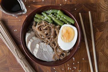 Asian food ramen noodles with meat, vegetables and egg in a brown bowl on a wooden board, soy sauce in a gravy boat, chopsticks, raw noodles