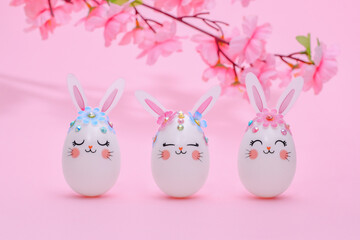 Three Easter eggs of a hare on a pink background with a flowering branch of an apple tree on the backdrop. White eggs decorated with pink rabbit ears, flowers and muzzles of rabbits Easter composition