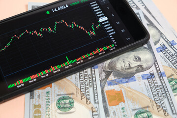 Stock exchange chart on smartphone screen and hundred dollar bills on beige background.