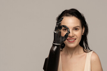 Beauty disabled Woman showing OK by prosthetic arm, artificial hand isolated on beige background. Surprised. Natural Skin Makeup Fresh Spa Women's Cosmetic Portrait. Beauty variety. Copy space - obrazy, fototapety, plakaty
