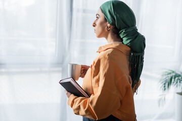 side view of arabian student with notebook and tea cup looking away at home.