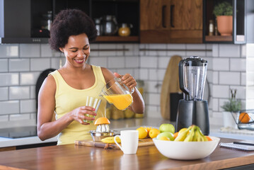 Young cheerful African-American woman drinking orange juice and surfing the internet in the kitchen at home after exercise