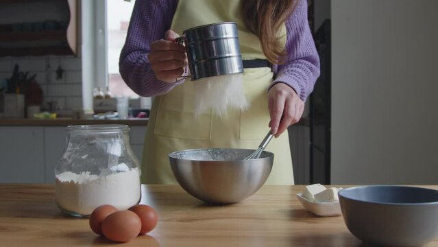 Woman in yellow apron is cooking at home, making dough, sifting the flour into bowl, using sifter cup, preferring homemade bakery, Slow motion.