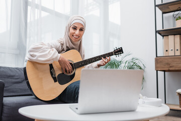 cheerful arabian woman looking at blurred laptop while learning to play guitar at home.