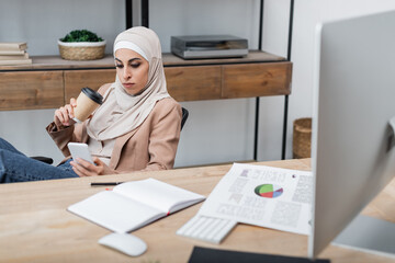 muslim woman with coffee to go and smartphone sitting near blank notebook and charts on desk.