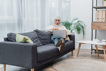 full length view of young muslim woman sitting on couch in living room and watching movie on laptop.