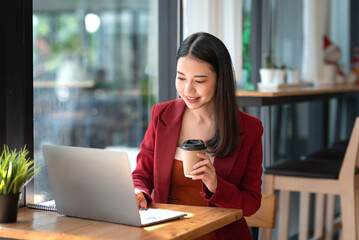 Confident beautiful Asian businesswoman typing laptop computer while holding coffee at café.