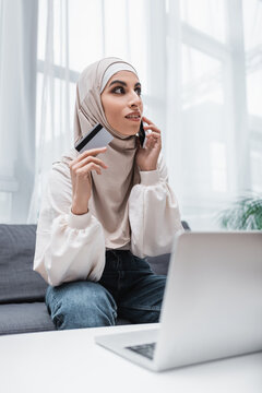 arabian woman holding credit card while talking on mobile phone near laptop.