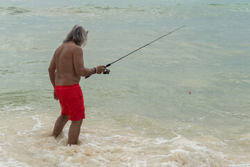 An elderly man with a naked torso is fishing in the sea. A man stands in the water and holds a fishing rod. Fishing in troubled waters.