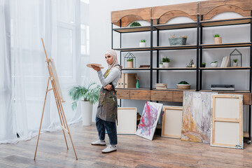 full length view of muslim woman in apron standing with palette near easel.
